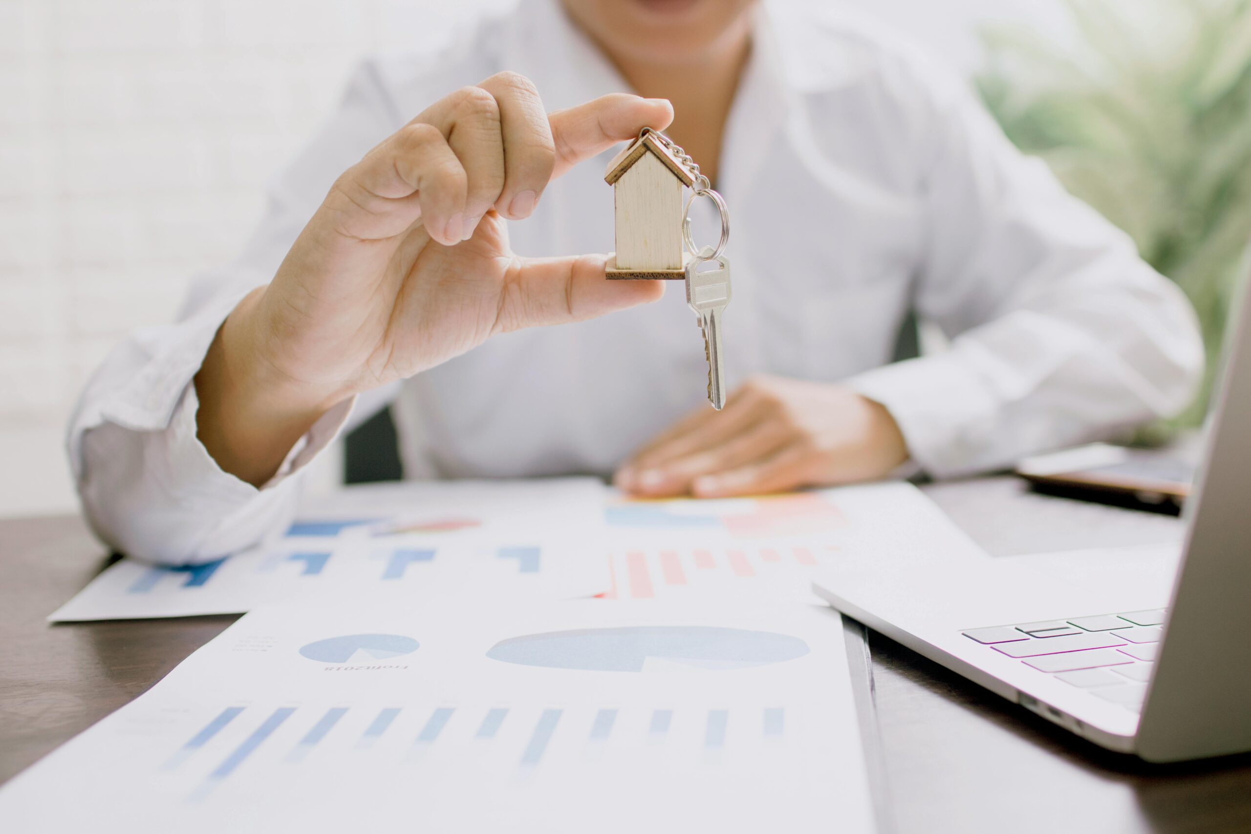 Businessperson holding a small wooden house keychain over financial documents and graphs, symbolising property investment and financial planning.