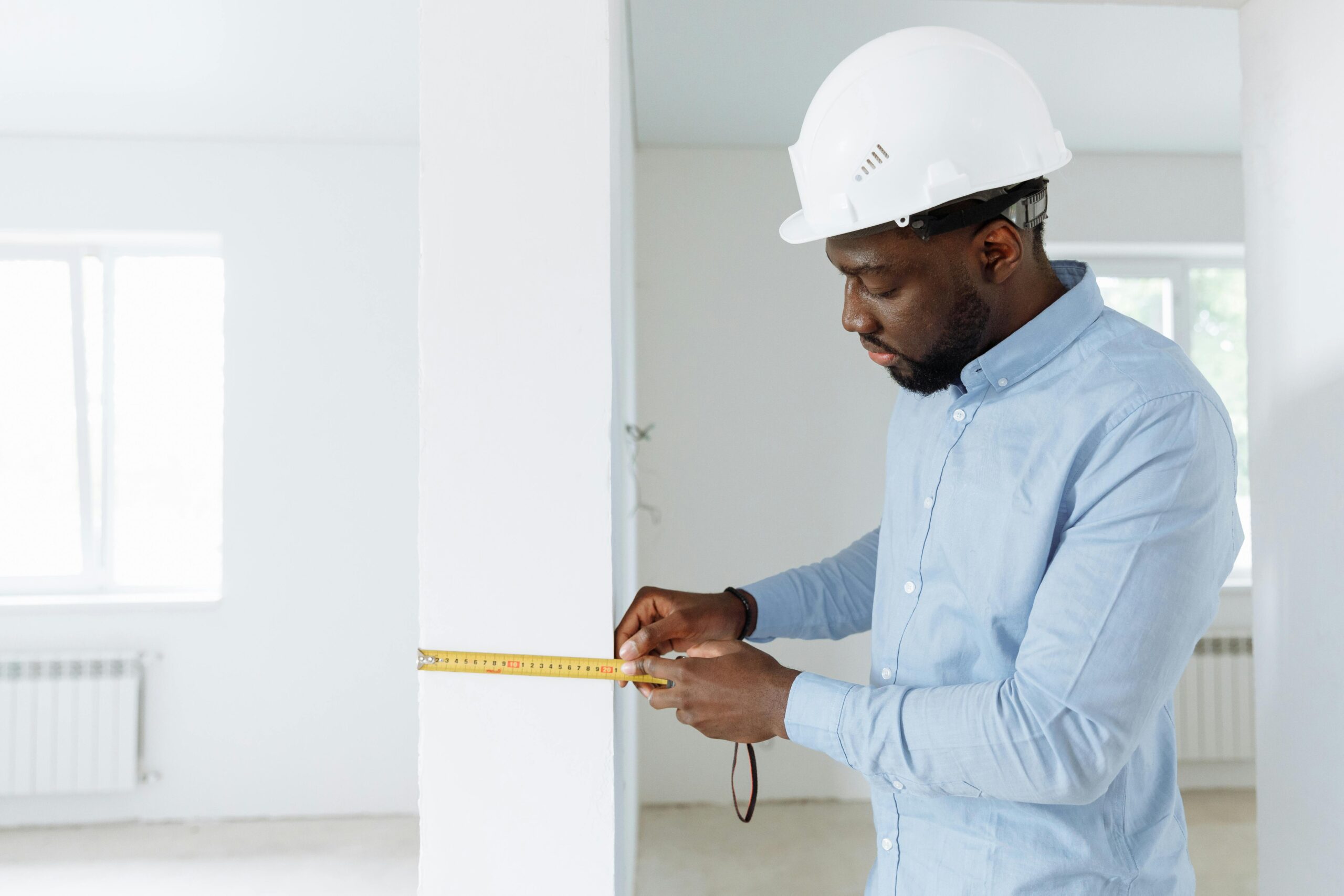 Construction professional in a hard hat measuring a wall with a tape measure inside a bright, unfinished building.